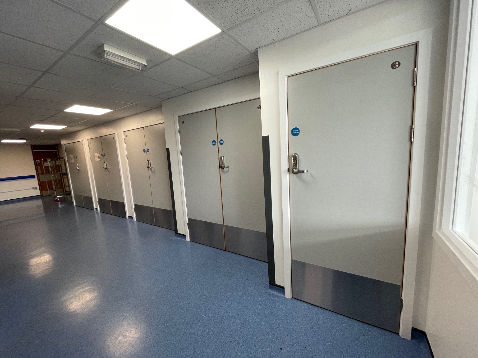 A corridor with blue flooring, white walls, and several closed doors with keypads and blue signage on each door. Ceiling lights illuminate the corridor.