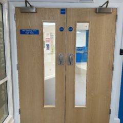 Double wooden doors with vertical windows, silver handles, and signs reading "Hot Desking Area Staff Only." A corridor and noticeboard are visible through the glass panels.