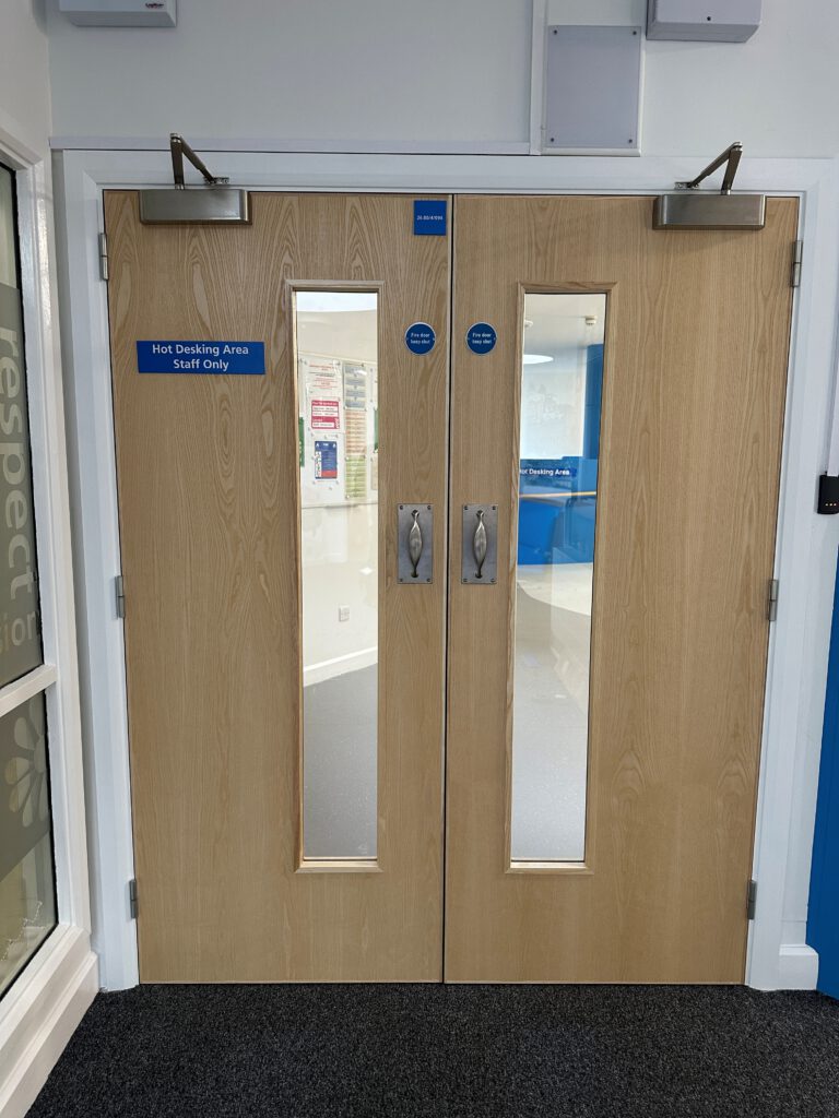 Double wooden doors with vertical windows, silver handles, and signs reading "Hot Desking Area Staff Only." A corridor and noticeboard are visible through the glass panels.