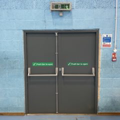 Double emergency exit doors with green "Push bar to open" signs, a fire alarm to the right, and an illuminated emergency exit sign above on a blue brick wall.