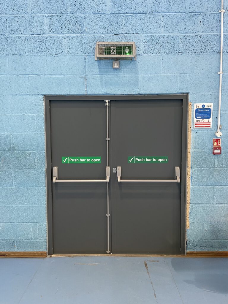 Double emergency exit doors with green "Push bar to open" signs, a fire alarm to the right, and an illuminated emergency exit sign above on a blue brick wall.