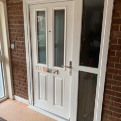 A white front door with a silver handle and letterbox, flanked by a tall glass panel, set in a brick wall with a tiled floor. House number 223 is visible on the letterbox.