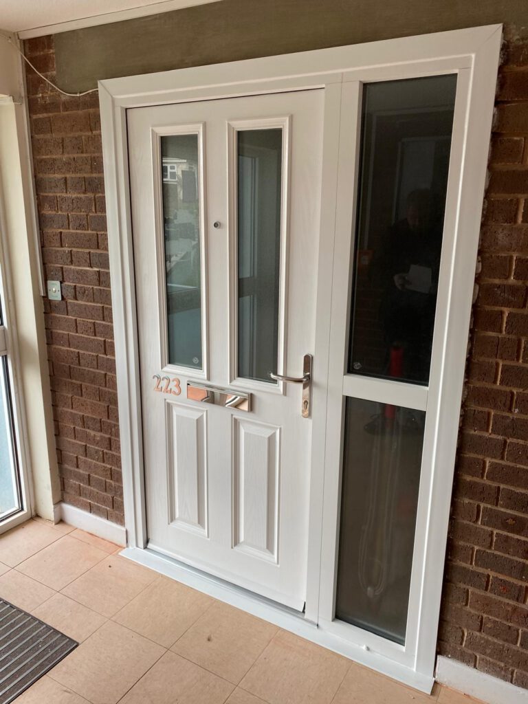 A white front door with a silver handle and letterbox, flanked by a tall glass panel, set in a brick wall with a tiled floor. House number 223 is visible on the letterbox.