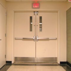 Double doors with push bars and narrow windows, an exit sign above, a fire extinguisher mounted on the right wall, and a single door on the left.