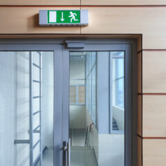 Glass double doors in a corridor with an illuminated green emergency exit sign above and a red fire alarm on the wall to the right.