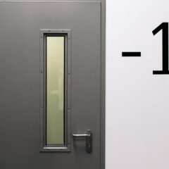 Grey door with a narrow vertical window and a metal handle next to a white wall displaying the black number “-1,” indicating a basement or underground floor.