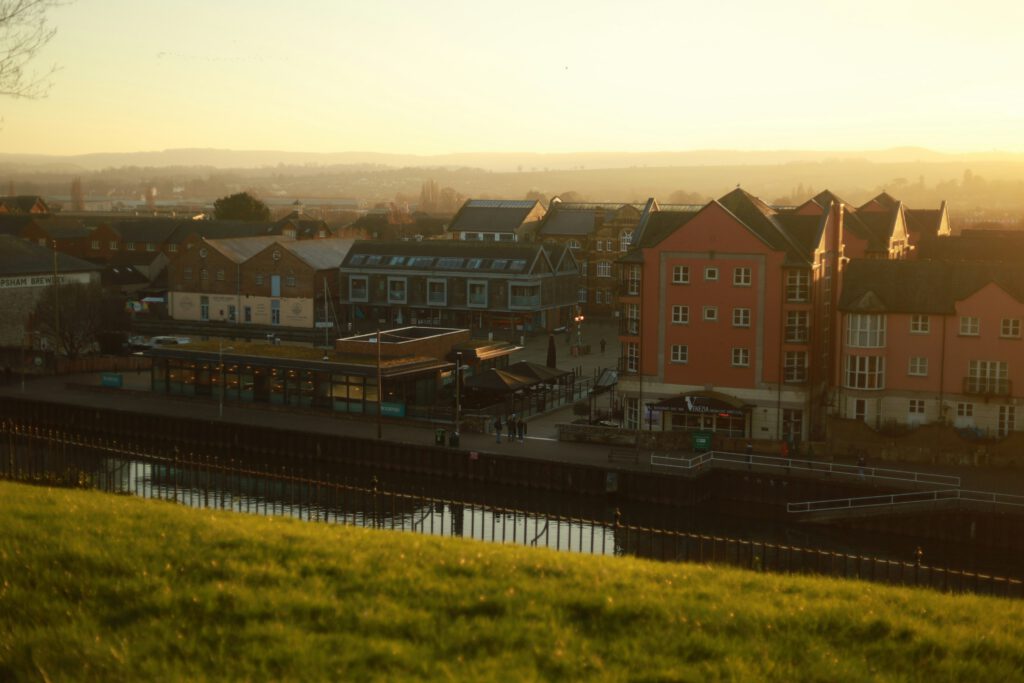 A view of several colourful buildings near a canal at sunset, with a grassy area in the foreground and hills visible in the background.